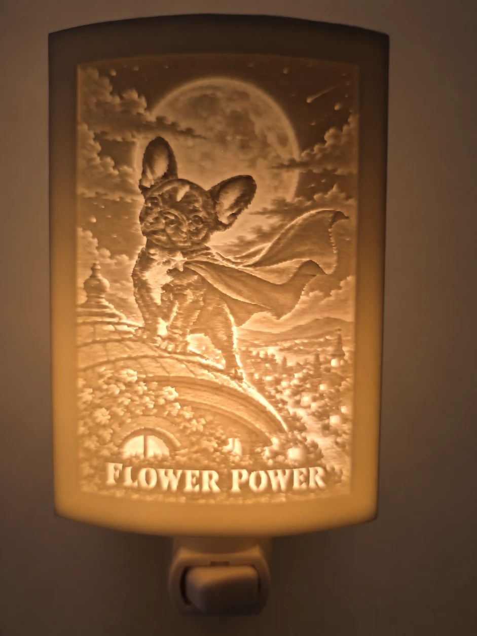 Physical nightlight prototype glowing warm amber: a curved panel showing Flower in a superhero cape above a moonlit bridge, with FLOWER POWER lettering and the lower switch housing visible. Background edited.